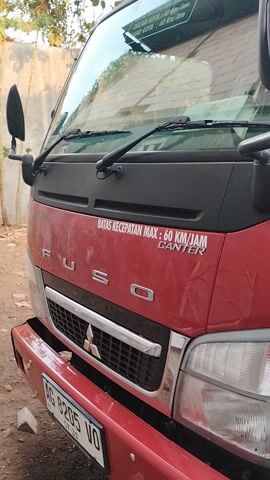 This is a close-up, rotating video of the front and side of a red Mitsubishi Fuso Canter truck parked outdoors. The truck is the primary subject. It is a commercial vehicle, likely used for hauling goods. The camera angle is at eye level with the truck's front, providing a clear view of its grille, headlights, and windshield. The truck appears to be in good condition, with a clean, shiny red paint job.

Visible text on the truck includes "FUSO" prominently displayed on the grille, along with the Mitsubishi logo. On the side of the truck, there are several white decals with red text. One decal reads "BATAS KECEPATAN MAX: 60 KM/JAM CANTER," indicating a maximum speed limit. Another decal says "DILARANG MEROKOK," meaning "No Smoking." There is also a rectangular sticker with a blue and yellow logo and the text "ELPIJI," suggesting it might be related to gas distribution. A license plate is partially visible, showing "AG 8205 VO".

The setting appears to be a yard or lot, possibly in Pehserut, Indonesia, as indicated by the location context. The ground is dirt or gravel. In the background, there are glimpses of trees and what might be a building or structure, possibly a covered area where the truck is parked. The lighting suggests it is daytime, with no strong shadows indicating harsh sunlight. There are no people visible in the video. The overall impression is a straightforward presentation of the truck and its branding.