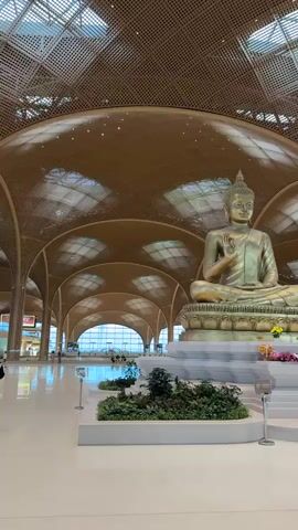 This video pans across a large, modern interior space, possibly an airport terminal, characterized by a striking vaulted ceiling made of wood panels with a geometric pattern. In the foreground, a large, golden Buddha statue sits in a meditative pose on a tiered platform adorned with greenery and flowers. Several people are visible in the midground and background, some standing in small groups, others walking. One person in a red shirt is standing near the Buddha statue, possibly taking a picture. The floor is a smooth, light-colored surface, and the overall impression is one of spaciousness and contemporary design, possibly with an Asian architectural influence due to the Buddha statue. The lighting appears to be artificial, possibly from the ceiling structure itself.