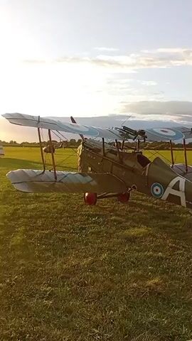 A biplane with the markings of the Royal Flying Corps is parked on a grassy field. The plane is olive green and has roundels on its wings and fuselage. The tail is painted with the French tricolor. A second biplane, this one with German markings, is parked behind it. The German plane has black crosses on its wings and fuselage. The sun is setting, casting long shadows across the field. The sky is a mix of blue and white clouds. The video appears to be shot from a low angle, looking up at the planes.