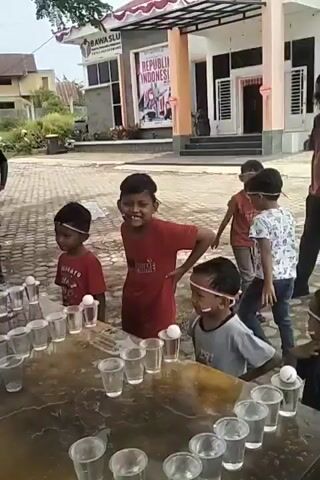A group of young boys are participating in an outdoor game. They are standing behind a table with several clear plastic cups lined up. Each cup contains a small white ball, and the boys are wearing headbands. The boy in the center, wearing a red shirt, is actively engaged in the game, with one hand extended. Another boy, to his right, also in a grey shirt, is focused on the cups. In the background, an adult male in a pink shirt and sunglasses observes the game. Further back, a building with "REPUBLIK INDONESIA" written on it is visible. The environment appears to be a public space, possibly a schoolyard or community area, with paved ground and some greenery.