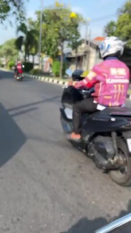 The video shows a person riding a black motorcycle on a paved road. The rider is wearing a white helmet and a brightly colored jacket with a design that includes pink, yellow, and black. They are also wearing black pants. The motorcycle has a visible license plate. In the background, there are trees with yellow flowers, other buildings, and another motorcycle being ridden by a person wearing a red shirt. The scene appears to be outdoors during the day, with sunlight casting shadows on the road. The camera angle is from another moving vehicle, suggesting the video was captured from behind the rider.