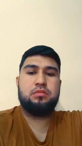 A close-up shot shows a man with dark hair and a beard, wearing a brown shirt. He is looking directly at the camera, his expression shifting slightly to a frown and then back to a neutral expression. He is indoors, standing in front of a plain, light-colored wall.