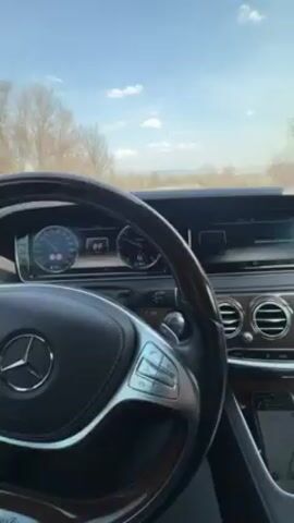 The video shows a first-person perspective from inside a Mercedes-Benz car. The driver is on a winding country road, with the speedometer reading around 90. The car has a wooden steering wheel, leather interior, and a modern dashboard. Outside, the weather is clear and sunny. There are bare trees lining the side of the road.