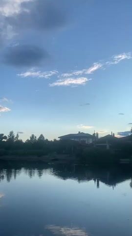 A static, vertical video shot at dusk, facing a calm body of water that reflects the sky and a tree-lined shore with houses. The sky is a clear blue with scattered clouds, some of which are dark and heavy, others wispy and illuminated by the setting sun. The water is smooth, creating a mirror-like reflection of the sky and the silhouettes of trees and buildings on the opposite bank. The overall impression is one of tranquility and peace as daylight fades.