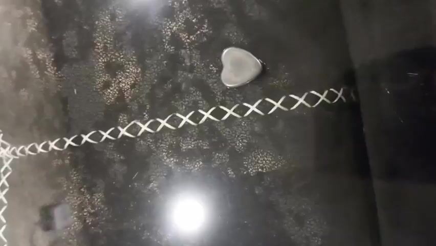 The video shows a dark, speckled surface with two heart-shaped metallic objects and a white braided cord. The cord is positioned horizontally across the frame, forming an X pattern. One heart-shaped object is located above and to the right of the cord, and another is below and slightly to the left of the first object. A hand with a thumb and index finger is shown reaching from the right side of the frame towards the upper heart-shaped object. The lighting suggests an indoor environment with overhead light sources reflecting off the surface.