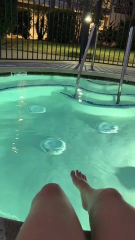 The video begins with a close-up view of a person's legs and feet submerged in a brightly lit, turquoise hot tub. Bubbles rise from the jets in the tub, creating ripples on the water's surface. Metal handrails are visible, leading out of the water. As the camera pans up and slightly to the left, it reveals a black metal fence surrounding the hot tub area. Beyond the fence, there are palm trees and a building with balconies. The sky appears to be dusk or early evening. The camera continues to pan upwards, showing more of the building and the sky. The sound of bubbling water and distant, indistinct voices can be heard. The video concludes with a wider shot of the hot tub and its surroundings.