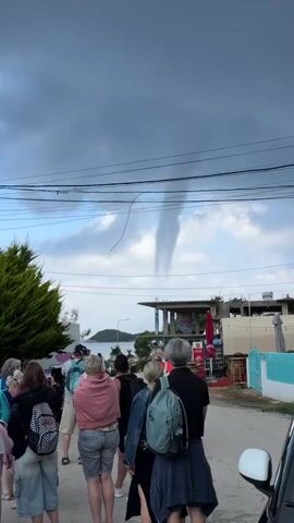 A group of people stands on a paved road, looking up and watching a waterspout extending from a cloud towards the sea in the distance. The waterspout is a prominent, funnel-shaped cloud column descending from a dark, cloudy sky.

The scene is a coastal area, with the sea and what appear to be islands or hills visible on the horizon. Several buildings, some appearing residential or commercial (one with red umbrellas suggesting a cafe), line the street. Overhead power lines stretch across the upper part of the frame, with one loose wire hanging down prominently. Green foliage is visible on the left and in the background. A white car is partially visible on the right side of the frame.

The people, dressed in casual clothing, are observing the weather phenomenon, with some holding up phones as if recording. The sky is largely overcast, though some patches of lighter sky are visible beneath the dark clouds. The time of day appears to be daytime. Graffiti, possibly reading "CRFT," is visible on one of the buildings.