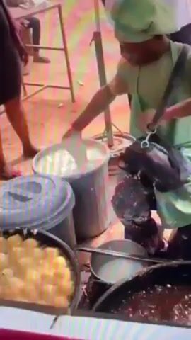 A person wearing a green hat and apron is scooping a white liquid from a large gray bucket into a metal container. The metal container is sitting among other containers holding what appears to be food. A person in dark clothing stands behind the food preparation area, and part of a white ladder is visible in the background.