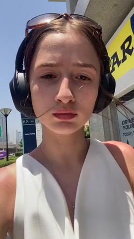 The video shows a young woman outdoors on a sunny day. She has light brown hair and wears headphones, a white top, and cream-colored shorts. She also has a small beige handbag and sunglasses. She holds the camera and takes a selfie video, rotating the camera to show her outfit. In the background are buildings and a person wearing an orange uniform. The location appears to be an urban area.