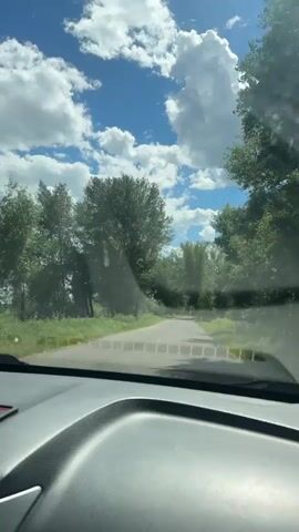The video captures a first-person perspective from inside a car driving down a paved road. The road is flanked by lush green trees on both sides, with a narrow strip of grass between the road and the trees. The sky is a vibrant blue with fluffy white clouds, indicating fair weather. The car's dashboard is visible in the foreground, partially obscuring the view of the road ahead. The road itself appears to be a rural or suburban route, with no other vehicles or pedestrians immediately visible. The scene conveys a sense of travel through a natural, tree-lined environment on a clear day.