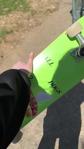 A person holds a bright green skateboard with the words “ALL DOGS” printed on it, followed by a graphic of a hotdog with wings. The back of the skateboard is black and a thin pink stripe runs between the black surface and the green underside. The person holding the board is wearing a black long-sleeved shirt. The video is shot outside on a sunny day on a gravel path with grass visible to the side. Another person wearing blue jeans and brown boots walks by in the background.