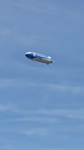 A blimp floats in a clear blue sky with a few wispy clouds. The blimp is primarily white with a blue tip and a blue stripe running along its length. Text and logos are visible on the blimp, including "ZF Next Generation Mobility" and a red "Fielmann" logo. The blimp is oriented with its nose slightly upward and appears to be moving from left to right across the frame. The lighting suggests it is daytime.