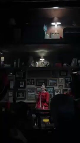 A dimly lit bar features a woman in a red dress singing into a microphone at a small table.  Behind her, on the upper part of the wall, is a shelf cluttered with objects.  Below the shelf, a collection of framed photographs covers the wall.  A few musicians stand behind her, one with a guitar.  A small, dim audience watches from the dark.  The lighting comes from a small lamp above the stage area.