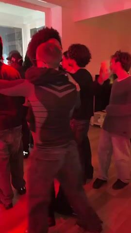 The video shows a group of people dancing and interacting in a room with red lighting. One man with an afro embraces another man. Many are wearing dark clothing and jeans. A few are holding drinks. The environment appears to be an informal gathering, possibly a party. There is a window and a television in the background.