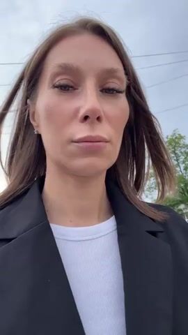 A medium shot shows a woman with brown hair and fair skin looking at the camera. She is wearing a black blazer over a white shirt. Her hair is windswept, suggesting she is outdoors. The background is slightly blurred but shows a building with windows and some trees. The sky appears overcast. The woman's expression is neutral.