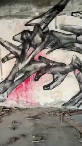 The video presents a close-up view of a graffiti mural on a distressed wall. The mural is predominantly black and gray, featuring abstract forms reminiscent of distorted hands or branches interweaving with each other. A splash of pink and orange hues is visible towards the lower center of the mural, adding a focal point of color. The wall shows signs of wear and tear, with patches of exposed concrete and peeling paint. In the foreground, the floor is covered with debris, including splintered wood and fragments of plaster, suggesting that the space is abandoned or under renovation. The camera slowly pans across the mural, offering different angles of the artwork and its integration with the dilapidated surroundings.
