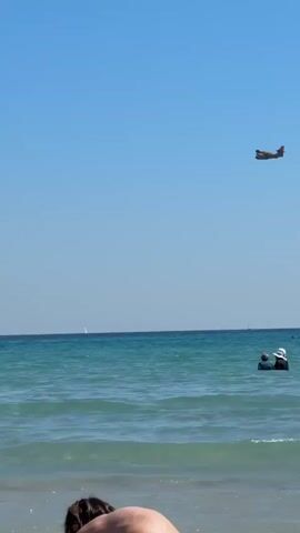 The video shows a bright, sunny day at a beach, likely in La Ciotat, France. The scene features a clear blue sky and calm, turquoise waters. In the foreground, a person's back and head are visible on the sandy shore. Further out in the sea, several individuals are swimming or wading, enjoying the water. Small, gentle waves break near the shoreline.

Dominating the upper part of the frame is a firefighting aircraft, resembling a Canadair CL-215 or CL-415, flying across the sky from right to left. The plane's presence suggests a potential wildfire operation, but the beachgoers below appear relaxed and unconcerned. In the distant background, gentle hills or mountains line the horizon, and faint outlines of small sailboats can be seen on the water. The overall impression is one of a serene summer beach day, with an unexpected but perhaps common sight of a firefighting plane passing overhead.