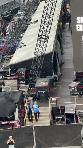 This video shows an aerial view of a large event setup, likely a concert or show, with a stage structure partially covered by a white tarp. Several pieces of stage equipment and trussing are visible. In the foreground, two individuals wearing blue vests and lanyards are walking up a set of stairs. One person is holding a piece of paper. To the right, a large screen displays text, partially obscured by the tarp, with the words "SHOW" and "EXC" visible. The environment appears to be an indoor arena or stadium, with seating visible in the background and a crowd of people. Various black equipment cases and carts are stacked around the stage area.