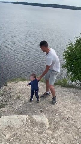 A man holds a young child's hand while standing on a rocky cliff overlooking a vast body of water. The man wears a white t-shirt and light shorts, carefully guiding the child, who is dressed in a dark blue outfit and sneakers, as they walk near the edge. The wide body of water, likely a river, is characterized by ripples on its surface. A small motorboat is visible on the water, creating a noticeable wake, and a larger vessel can be seen further in the distance.

In the background, a distant city skyline and a long bridge stretch across the horizon. The sky is partly cloudy, with patches of blue sky and fluffy white and grey clouds. Green bushes and evergreen trees grow on the steep slope leading down to the water. The scene takes place during daytime, with clear visibility. The location, identified as Пятый километр, Russia, suggests a viewpoint over a large river like the Volga. No visible text is present in the video.