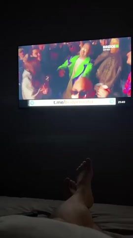 The video shows a person lying in bed, watching a television screen. The television displays a broadcast from Bridge TV, a Russian music channel. The screen shows a music video or live performance featuring several people, some of whom appear to be singing or dancing. The overall mood of the performance seems energetic and celebratory, with colorful costumes and bright lighting.

The on-screen text indicates the time is 21:40 and shows the channel logo "BRIDGE ФРЕШ" (Bridge Fresh). There is also a Telegram channel URL visible: t.me/bridgemedia. The setting appears to be a dimly lit room, likely a bedroom, given the presence of a bed and the feet of the person filming in the foreground. The footage captures a moment of leisure, with the viewer relaxing while enjoying entertainment. The video has a casual, home-video feel.
