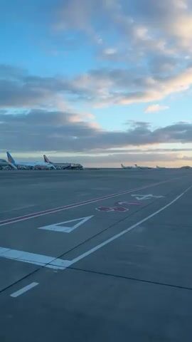 The video presents an exterior view of an airport runway under a blue sky with scattered clouds. The ground surface is made of gray concrete with painted white and pink markings. In the distance, several airplanes are parked. The light suggests it is either early morning or late afternoon. No people are clearly visible in the frame.
