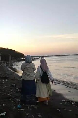 Two individuals wearing hijabs and long skirts walk along a dark, trash-strewn beach. The individual on the left wears a light-colored jacket over a blue skirt, and the individual on the right wears a light-colored top with a patterned skirt and a black backpack. They are walking away from the camera towards the water, where a faint sunset casts a warm glow. The water is calm, and in the distance, there are small structures and boats. The beach is littered with debris, including plastic bottles and wrappers.