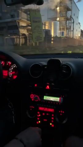 The video shows the interior of a car at dusk, with the camera pointing forward through the windshield. The dashboard is visible, with red illuminated gauges and a radio displaying the word "ROCK". The driver's hand is visible on the gear shift. Outside, a road is visible with industrial buildings and trees. The sky is a mix of orange and blue, indicating sunset. The car appears to be in motion.