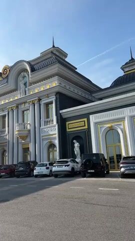 The video captures an exterior view of a large, ornate building with neoclassical or Baroque revival architecture. The building features a grey and white facade with extensive gold detailing on columns, cornices, window frames, and decorative elements. Statues of female figures are visible on the roofline and a lower section of the building. A prominent golden, arched canopy extends from the building's entrance area. Several cars, including sedans and SUVs of various colors (black, white, red, silver), are parked on an asphalt lot in front of the building. The sky is clear and blue with minimal clouds. The camera slowly pans from right to left, revealing more of the building's facade and the parked vehicles.