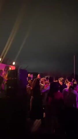 The video depicts a nighttime outdoor gathering with a crowd of people.  Several spotlights or beams of light cut through the dark, cloudy sky, originating from the left side of the frame and pointing upwards and to the right. In the foreground, the silhouettes of many individuals are visible, suggesting they are standing close together. Some appear to be dancing or moving within the crowd. On the left side of the image, a speaker or sound system is partially visible, indicated by a glowing orange light. The overall atmosphere appears to be a concert or party.