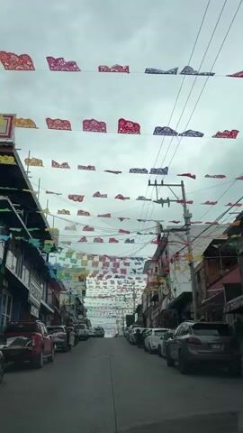 The video shows a street lined with buildings on both sides. The street is paved and appears to be a public road. Several cars are parked along the side of the street. A few people are visible walking on the sidewalk. The sky is overcast. The most prominent feature of the scene is the numerous strings of colorful paper banners, or papel picado, strung across the street between the buildings, creating a festive atmosphere. There are also utility poles and wires present. The camera appears to be positioned inside a vehicle, looking forward down the street.