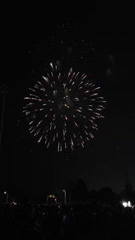 The video captures a fireworks display at night. The camera is positioned low, looking up at the dark sky filled with exploding fireworks. The fireworks are predominantly red and white, bursting in large, radial patterns. Silhouetted figures of people are visible in the foreground, appearing to be gathered in an outdoor space, possibly a park or field, watching the display. Lights from streetlights and distant buildings are faintly visible near the horizon line, along with the dark shapes of trees. The scene suggests a celebratory event taking place after dark.