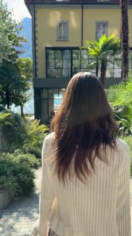 A woman with long, dark hair walks away from the camera on a cobblestone path. She wears a white, cropped sweater with flared sleeves and a short, floral print skirt. She carries a dark clutch in her right hand and has her left arm bent at the elbow. She is walking towards a light yellow building with large, dark-framed windows. Palm trees are visible on the right side of the building. To the left of the path, there is lush greenery, including ferns and bushes. Beyond the greenery and to the left of the building, a body of water is visible, with mountains in the distance. The sun is casting shadows on the path.