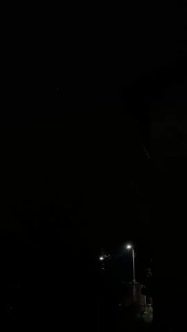 A nighttime video captures a distant lightning strike illuminating the dark sky for a fraction of a second. The lightning appears as a faint, white flash. In the foreground, a streetlamp is visible, casting a warm glow on its surroundings. Trees and a building silhouetted against the dark sky are also present. The lightning is brief and does not appear to be close to the camera.