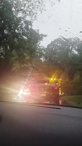 The video captures a view from inside a car, looking out through a rain-streaked windshield. The road ahead is lined with trees on both sides, suggesting a rural or wooded area. Traffic is visible, with brake lights of other vehicles illuminated. Several emergency vehicles, identified by flashing red and blue lights, are present. The weather appears to be inclement, with rain actively falling and affecting visibility. The overall scene suggests a traffic incident or a situation requiring emergency response on a wet, overcast day.
