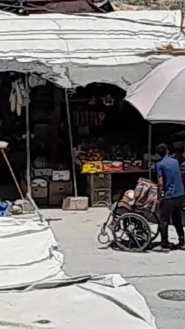 A man in a blue shirt pushes a wheelchair loaded with belongings down a street in front of a market stall. Another man in a light brown jacket stands by the stall, and a child in a pink shirt runs nearby. The market stall has a white awning and displays produce.