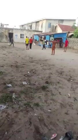 Several people are playing soccer on a dirt field in what appears to be a residential area. There are buildings visible in the background. Some people are watching the game from near a small, blue-painted structure. Several pairs of sandals are scattered on the ground. One player wearing an orange shirt dribbles the ball towards the camera.