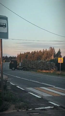 A truck carrying many round bales of hay drives along a road. To the left of the road is a bus stop sign and a pedestrian crossing. To the right of the road is a yellow diamond shaped sign with a pedestrian symbol. The sky is cloudy and the sun appears to be setting.