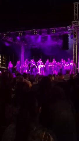 A large group of people are on a stage performing. They are in front of a black backdrop with many lights shining on them. The lights are white and purple and create beams that go out into the darkness. The group consists of men and women dressed in black clothing with some wearing white. In front of the stage, a large audience is watching the performance. The audience members are mostly facing away from the camera, looking towards the stage. The scene appears to be outdoors at night, as indicated by the darkness surrounding the lit stage and audience.