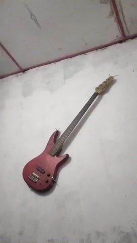 A red electric bass guitar is mounted on a white wall in a vertical, slightly angled position. The guitar has a light-colored headstock, a dark fretboard, and two pickups on its body. There are red painted details on the ceiling above the wall. The camera remains static throughout the video.