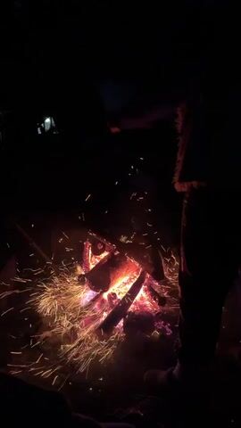 The video depicts a campfire burning in the dark. The flames are predominantly red and orange, with some hints of blue at their base. Embers are flying out from the fire. A person's silhouette stands near the fire, partially obscuring the view of the flames. The environment surrounding the fire is dark. In the background, faint lights suggest a building or other structure.