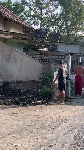 The video shows two men in an outdoor setting, likely a residential area. The man in the foreground is wearing a dark t-shirt and shorts, and is standing next to a large water cooler bottle. The man in the background is shirtless and wearing a red sarong. He appears to be holding something in his hand. They are standing near a building with a tiled roof and a fence. The ground is dirt and gravel. The lighting suggests it is daytime.