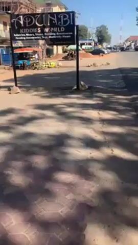 The video shows a street scene with a sign for "Adunbi Kiddies World" selling various baby and maternity items. In the background, there's a street vendor selling produce, possibly watermelon, under a canopy. Cars pass by in the distance. The camera appears to be held stationary. The day is bright and sunny, casting shadows on the ground. Sounds of a vehicle horn are heard.
