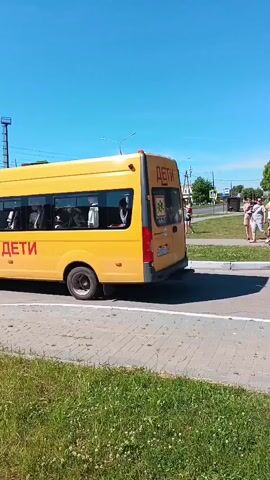 The video opens with a yellow bus, with the word "ДЕТИ" (children) written in red on its side, driving from left to right across the frame. Another yellow bus, also marked "ДЕТИ", follows shortly after. People are gathered on a grassy median on the right side of the road, watching the buses. A man in a blue t-shirt and grey shorts is visible in the foreground on the right. The sky is clear and blue, and there are trees in the background.