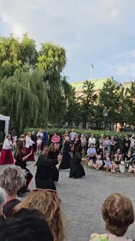 A group of dancers in black dresses are performing a synchronized routine in an outdoor, open-air venue. They are positioned in a semi-circle with the audience seated in front of them, some on the ground and others on chairs. Behind the dancers, a large weeping willow tree is visible, with lush green foliage. Further back, a white tent is set up, and beyond that, a building with a green roof and a flag is present. The sky is clear and blue. The dancers are moving their arms and bodies in unison, their faces focused on their performance. Some audience members are watching attentively, while others are talking amongst themselves. The atmosphere appears to be that of a cultural event or festival.