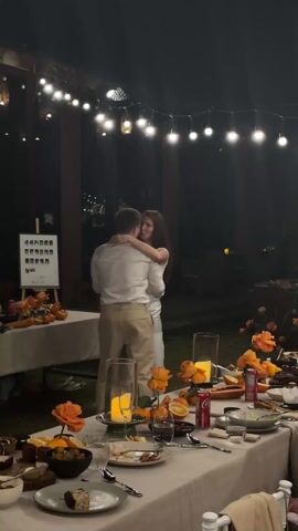 At an outdoor evening event with string lights, a man and a woman wearing light-colored clothes are dancing together closely between two long tables. The man has short hair and is wearing a white shirt and tan pants. The woman has long brown hair and is wearing a white dress with a slit. Tables are decorated with orange flowers, fruit, candles, and food items. There is a small white board with pictures on it. Someone can be seen sitting at one of the tables to the left.