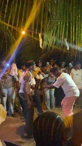 The video shows a group of people dancing in an outdoor setting, likely at night. The area is decorated with hanging lights and palm leaves, creating a festive atmosphere. Two men are in the center, engaging in enthusiastic dance moves. Others surround them, watching, clapping, and recording with their phones. Some are wearing casual clothing while others have uniforms. The lighting is a mix of bright artificial lights and natural darkness. The ground appears to be sandy or dirt. There is music playing. The scene conveys a sense of celebration and fun.