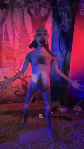 A creepy scene unfolds in a room illuminated with red, blue, and purple lighting. A grotesque statue of a naked, emaciated figure with green skin, black hair, and a long, curled tongue juts out. The statue stands with its arms outstretched and its face contorted in a menacing expression. Behind it, a painted backdrop depicts a dark forest with ghostly trees and a towering pagoda. The room features a pile of wood arranged like a pyre, with an artificial glow emanating from within. A human skull rests atop the woodpile. A person can be briefly seen in the left side of the frame. The scene creates a spooky and unsettling atmosphere.
