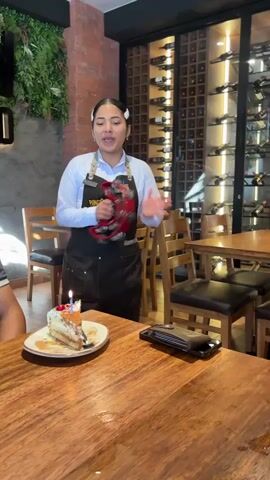 A restaurant with a woman in a server uniform standing at a table in front of a slice of cake, shaking a tambourine. A man in a similar uniform stands next to her, also shaking a tambourine. Behind the pair is a glass-enclosed wine cellar. The table has a black wallet and phone on it, indicating the location may be for a celebration.