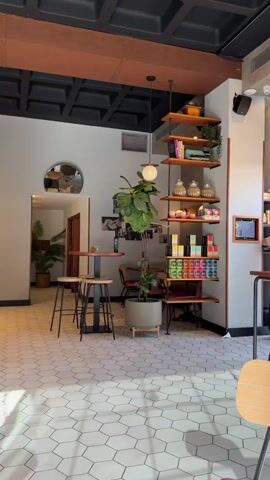 This media file captures the interior of a modern cafe in Pla Parcial Platja, Spain. The scene is empty of people, suggesting it might be early in the morning or before opening hours. The time of day is likely daytime, indicated by the bright sunlight casting long shadows across the hexagonal tiled floor. The weather appears to be pleasant, as suggested by the warm lighting and inviting atmosphere.

The cafe features a contemporary design with a black coffered ceiling and exposed copper accents. The walls are painted in a neutral light gray, creating a clean and airy feel. Dominating the space is a tall, leafy potted plant, adding a touch of nature to the urban setting.

Prominently displayed is a wooden shelving unit filled with various items. These include books, decorative glass jars, teacups, and colorful boxes of what appear to be tea or coffee. A vintage typewriter sits atop the shelves, adding a retro touch to the decor.

To the left, a wooden counter with a coffee machine is visible, hinting at the cafe's primary function. Opposite the counter, a small round table with two stools stands near an arched doorway, suggesting a potential seating area. Further into the cafe, other tables and chairs can be seen, some with a red hue.

A decorative circular mirror hangs on the wall, reflecting a glimpse of the interior. Artwork or photographs are also displayed on the wall, adding to the cafe's aesthetic. The overall impression is a stylish and welcoming space, designed for relaxation and enjoyment of coffee or tea. There is no visible text within the media file.