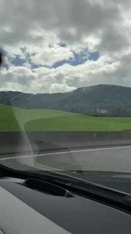 The video shows a view from inside a vehicle traveling on a highway. The road has multiple lanes separated by white lines, and a concrete barrier runs alongside the highway. Outside the vehicle, there is a lush green field adjacent to a wooded hillside. In the distance, rolling hills and mountains are visible under a partly cloudy sky. The interior of the vehicle is partially visible in the foreground, including a dashboard and part of the windshield. The scene appears to be captured during daylight hours.